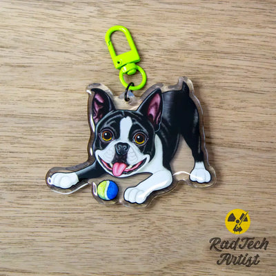 Acrylic Boston Terrier keychain with epoxy finish, featuring a black and white tuxedo pup crouched low with a tennis ball. Attached to a neon green lobster clasp, displayed on a dark wood background with the Rad Tech Artist logo.