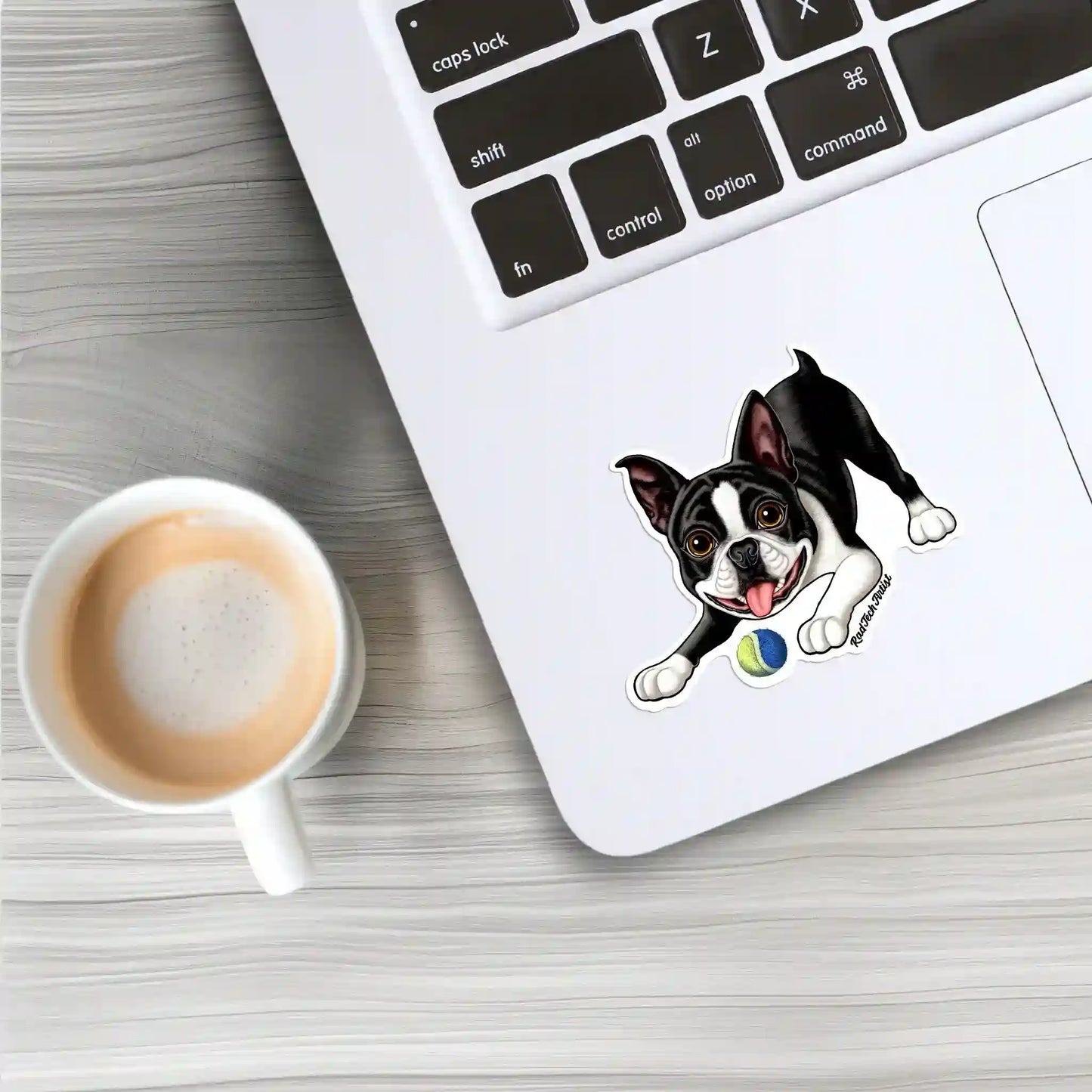 Mock-up image of a Boston Terrier vinyl sticker placed on a silver laptop near the keyboard, with a coffee cup beside it on a light wooden table. The sticker features a playful black-and-white Boston Terrier with a tennis ball.