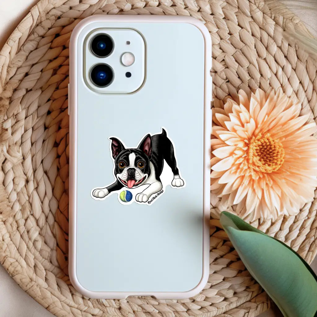 Photo of a tuxedo Boston Terrier sticker on the back of a white phone case. The playful black and white dog is shown with its tongue out and a yellow-and-blue tennis ball in front. The phone rests on a woven mat next to a peach flower and green leaf for a soft, styled look.