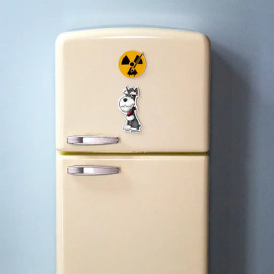 Actual photo of a Miniature Schnauzer magnet on a small cream-colored toy refrigerator. The magnet features a salt and pepper Schnauzer with a red collar and a grumpy expression. Above it is a round yellow Rad Tech Artist logo magnet with a radiation symbol and paintbrush.
