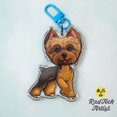 Acrylic Yorkshire Terrier keychain with epoxy finish, showing a blue and gold Yorkie standing proudly. Attached to a bright blue lobster clasp, displayed on a mint background with the Rad Tech Artist logo in the corner.