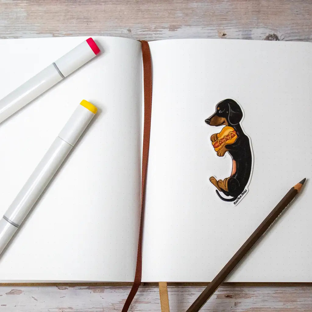 Photo of a black and tan Dachshund sticker holding a hot dog pillow, placed on an open sketchbook with markers and a pencil nearby on a wood-textured surface.