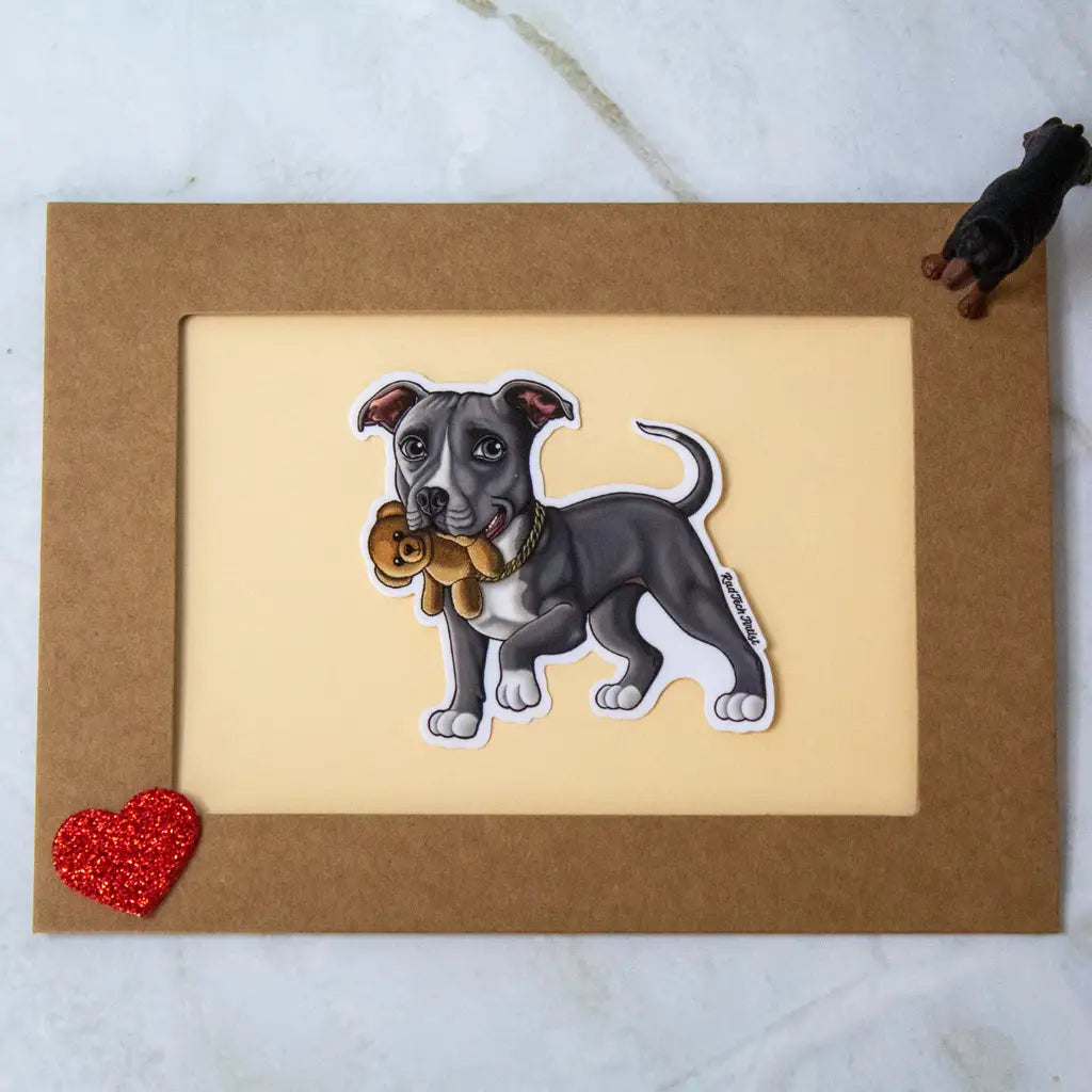 Actual photo of a blue American Pit Bull Terrier vinyl sticker placed on a bordered card. The pit bull has a sweet expression and is holding a teddy bear in its mouth. The card is decorated with a red glitter heart in the bottom left corner and a small black dog cutout in the top right.