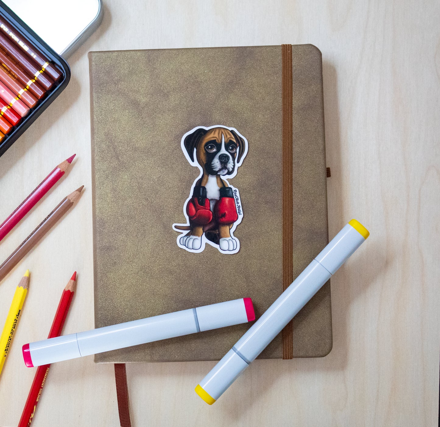 Boxer puppy vinyl sticker featuring a fawn and white pup wearing red boxing gloves, applied to a brown hardcover notebook. Surrounded by colorful markers and pencils on a light wood desk.