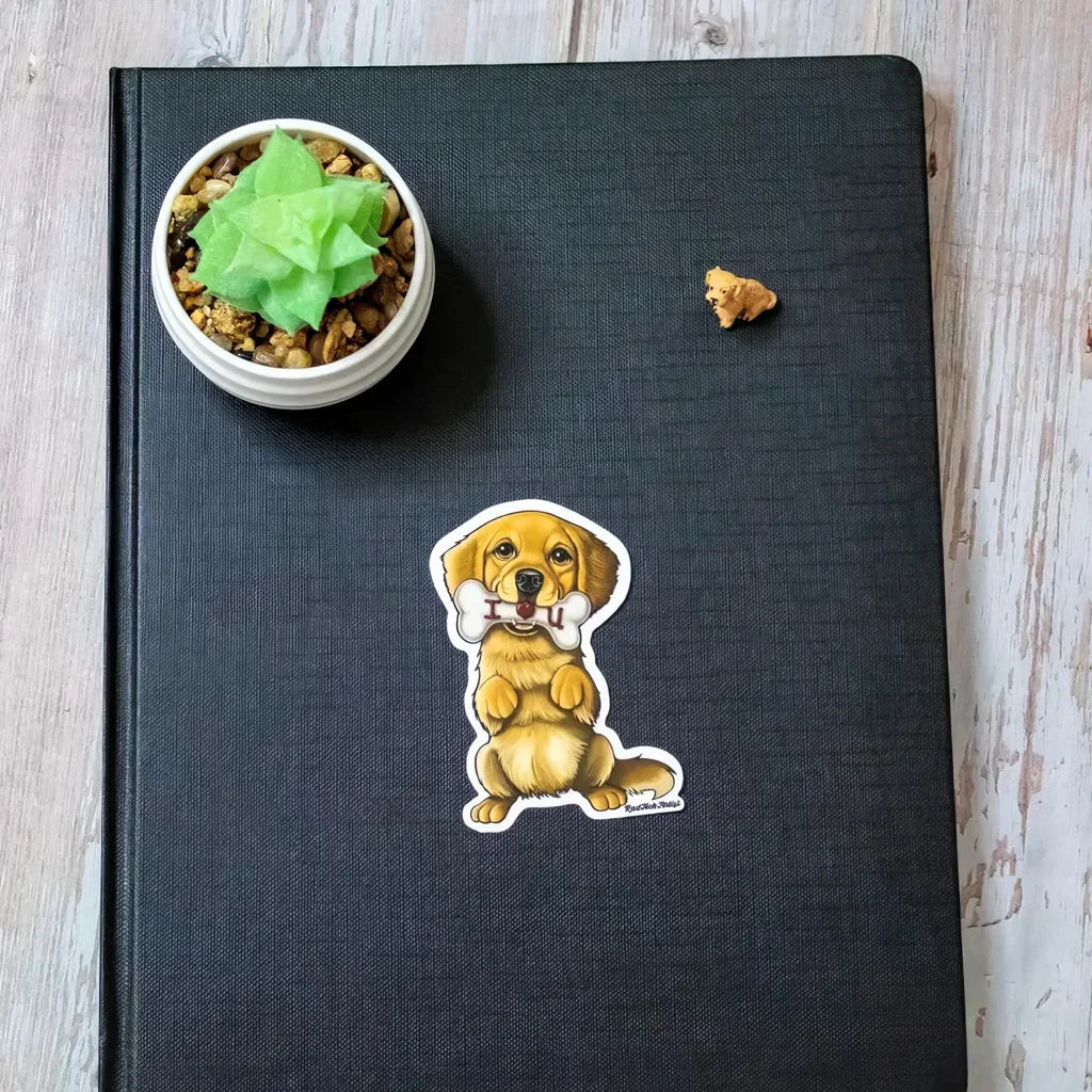 Actual photo of a Golden Retriever sticker placed on a dark textured notebook cover. The illustrated dog is holding a bone that says “I ❤️ U” and sits upright with a sweet expression. A small succulent and a treat are nearby on the wood surface.