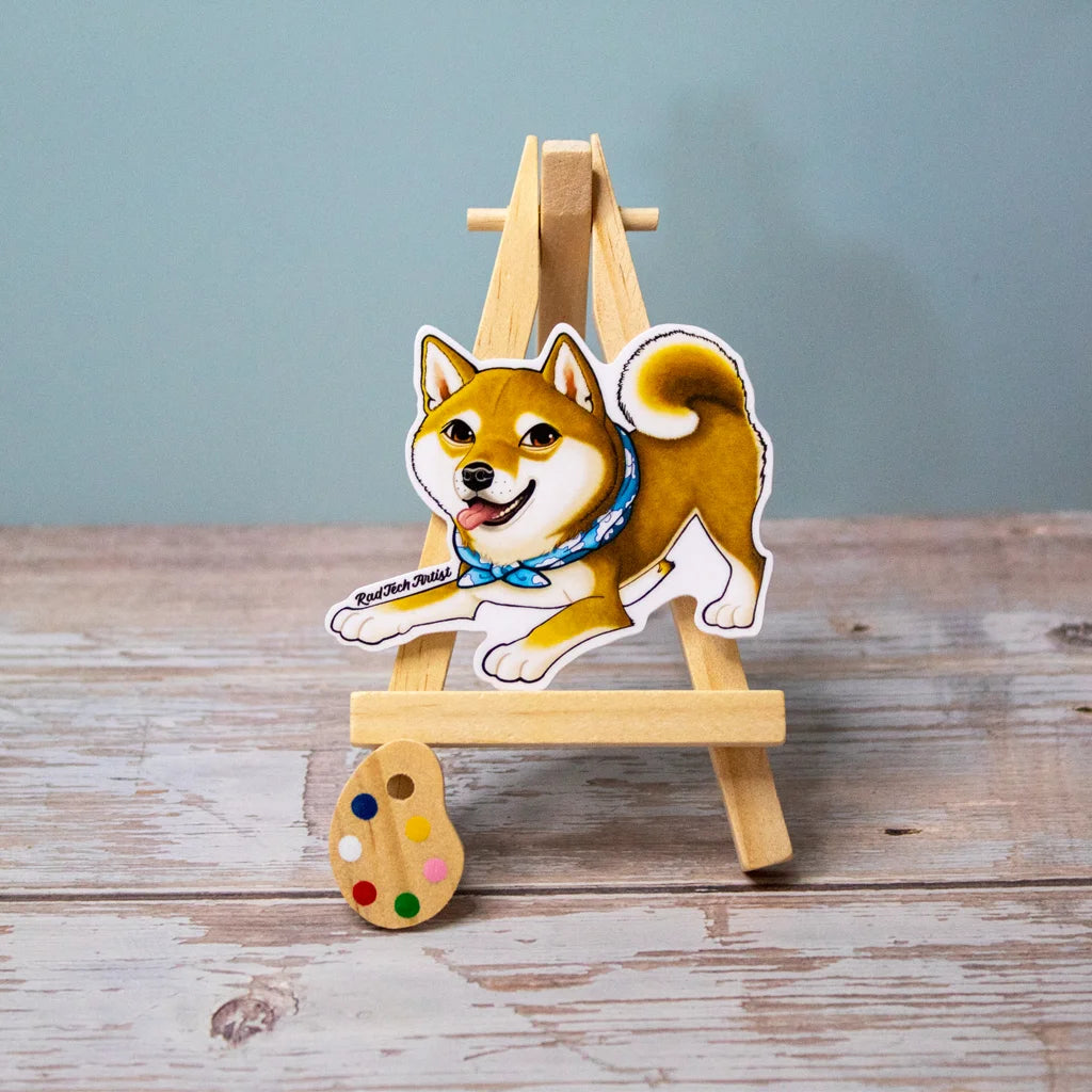 Vinyl sticker of a red Shiba Inu in a playful stance with a bright expression and curled tail, wearing a blue bandana, displayed on a wooden easel.