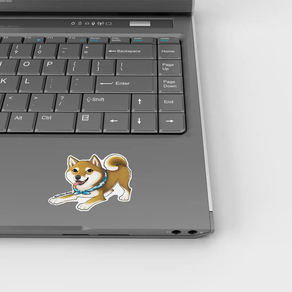 Mock-up of a red Shiba Inu vinyl sticker placed on a silver laptop, shown beneath the keyboard. The playful dog has a blue patterned bandana and is positioned in a crouching pose with a happy expression.