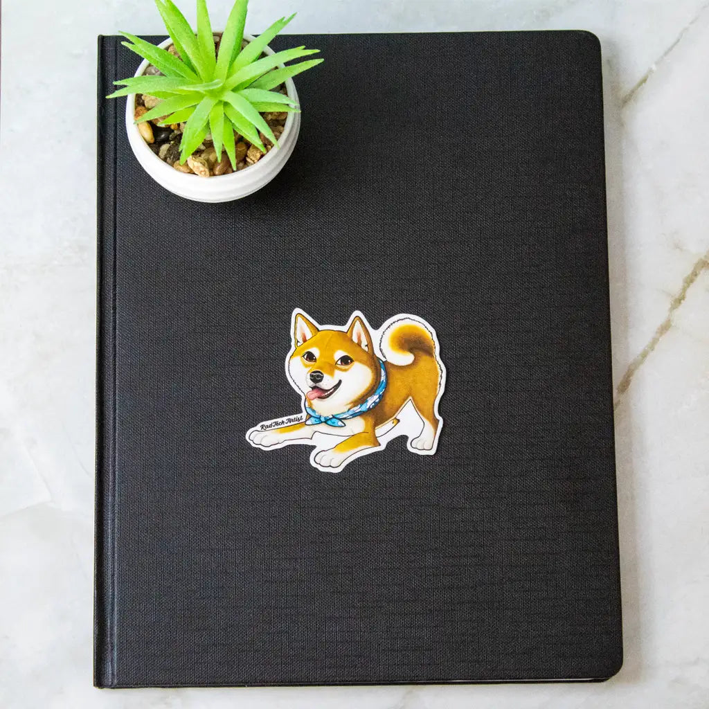 Actual photo of a red Shiba Inu vinyl sticker with a blue bandana, placed on a black textured notebook. A small potted succulent sits nearby on the marble surface.