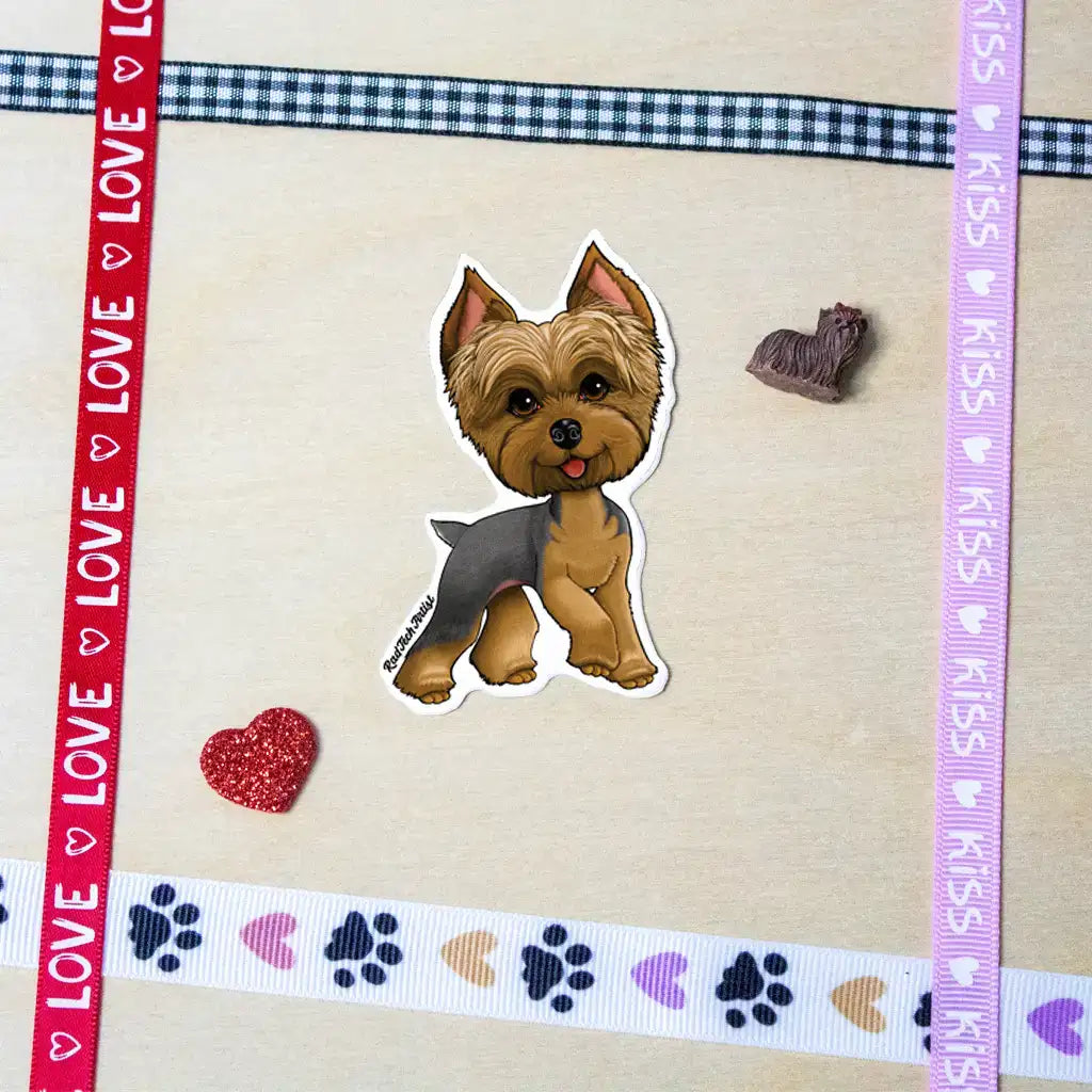 Photo of a blue and gold Yorkshire Terrier vinyl sticker with a sassy expression, placed on a beige surface decorated with colorful ribbon borders, paw prints, hearts, and a small Yorkie figurine.