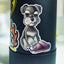 A close-up of a black insulated tumbler with a die-cut sticker of a salt and pepper schnauzer puppy sitting with a torn pink slipper. The tumbler is shown against a softly blurred light gray background.