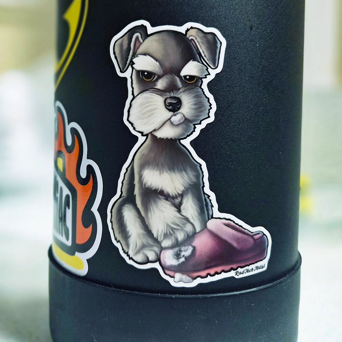 A close-up of a black insulated tumbler with a die-cut sticker of a salt and pepper schnauzer puppy sitting with a torn pink slipper. The tumbler is shown against a softly blurred light gray background.