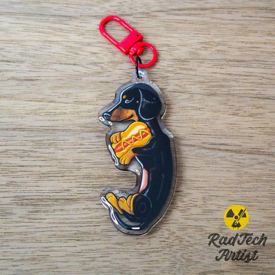 Acrylic black and tan Dachshund keychain with epoxy finish, featuring a playful pup holding a hot dog. Attached to a red lobster clasp, displayed on a warm wood background with the Rad Tech Artist logo.