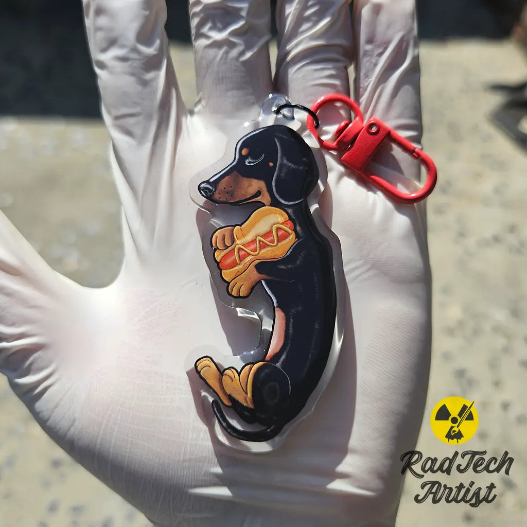 Black and tan Dachshund acrylic keychain with epoxy finish, featuring a playful pup holding a hot dog. Attached to a red lobster clasp and held in a gloved hand under sunlight, with the Rad Tech Artist logo in the corner.