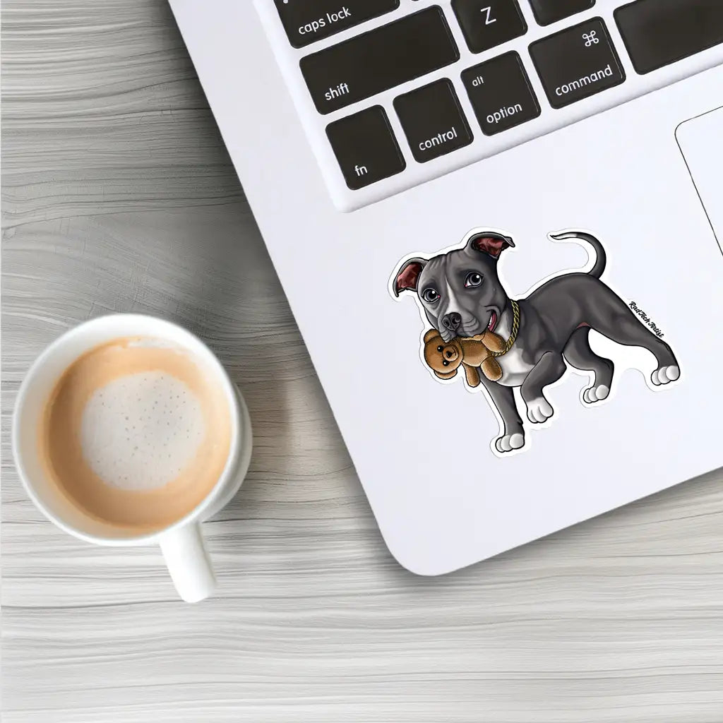 Mock-up image of a blue Pit Bull sticker on the back of a silver laptop. The dog has a gentle expression and is holding a small brown teddy bear in its mouth. The laptop sits on a light wood desk next to a coffee cup and part of a keyboard.