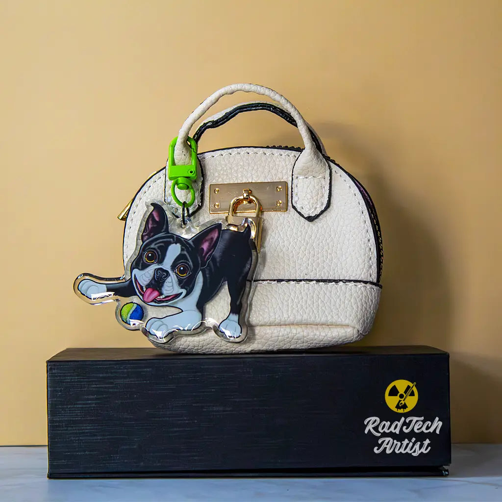 Acrylic Boston Terrier keychain with epoxy finish, showing a black-and-white dog in a playful bow pose with tongue out and a small tennis ball. Attached with a bright green lobster clasp to a textured white mini purse with gold lock hardware, displayed on a black box with the Rad Tech Artist logo.
