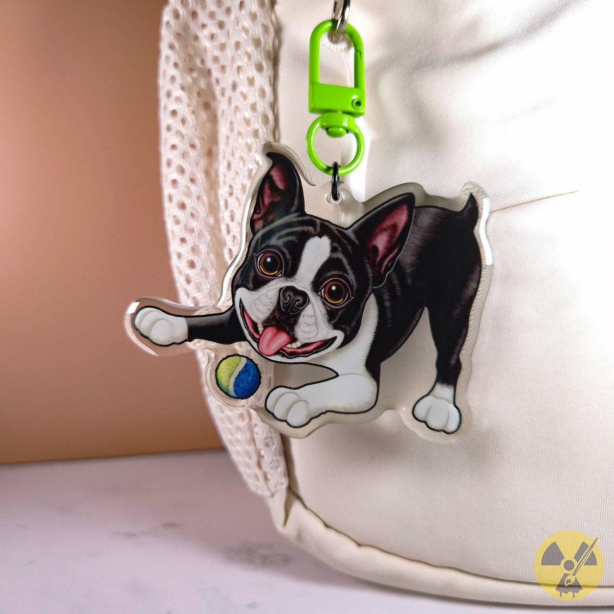 A custom die-cut acrylic keychain featuring a black and white Boston Terrier dog playing with a blue and yellow ball, attached to a bright green key clasp. The keychain is shown hanging next to a white textured fabric bag.