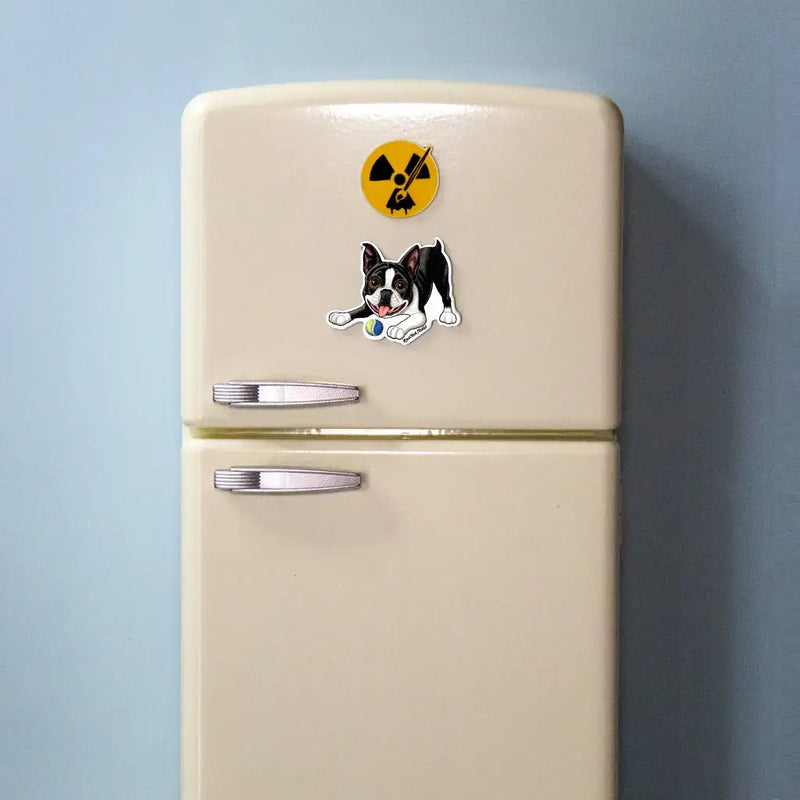 Actual photo of a black and white (tuxedo) Boston Terrier magnet on a small cream-colored toy refrigerator. The magnet features a happy Boston Terrier with its tongue out, playfully crouched with a yellow and blue tennis ball. Above it is a round yellow Rad Tech Artist logo magnet with a radiation symbol and paintbrush.