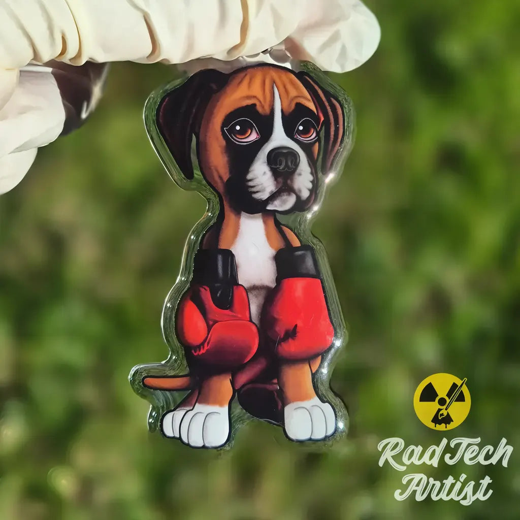 Hand holding a boxer puppy acrylic keychain with red boxing gloves and glossy epoxy finish against a green outdoor background.