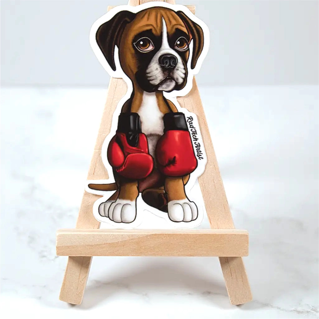 Sticker of a fawn boxer puppy wearing red boxing gloves on a small wooden easel against a white background