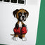 A die-cut sticker of a cute cartoon fawn  boxer puppy wearing red boxing gloves, placed on the silver surface of a laptop next to a green background.