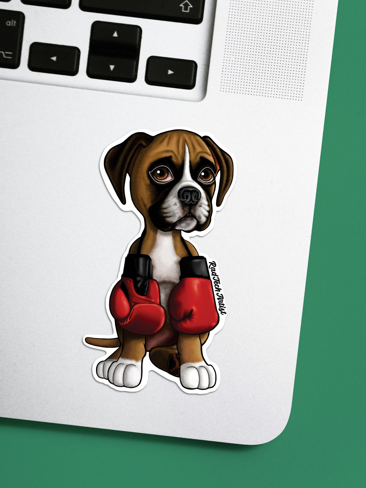 A die-cut sticker of a cute cartoon fawn  boxer puppy wearing red boxing gloves, placed on the silver surface of a laptop next to a green background.