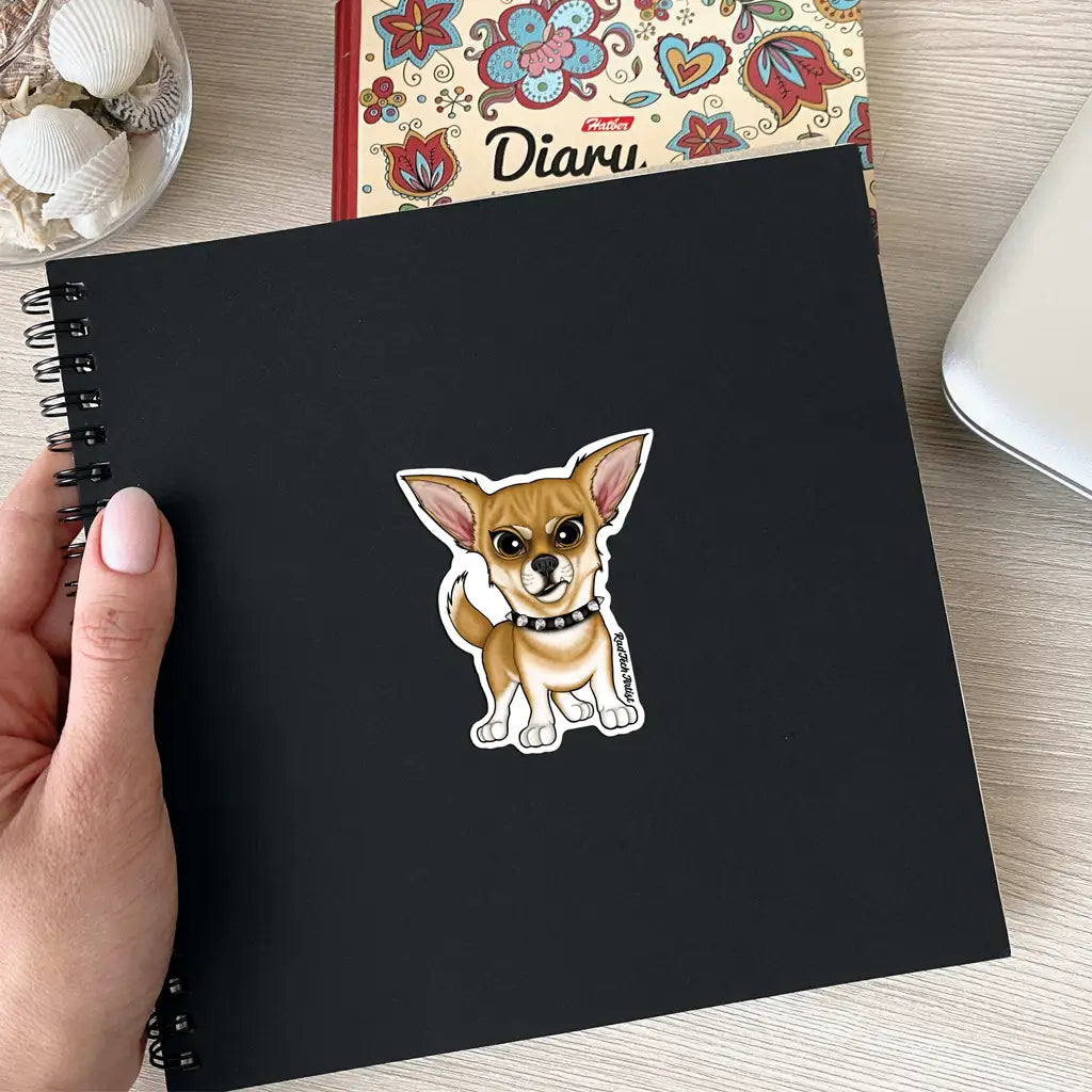Photo of a fawn-colored Chihuahua sticker placed on the cover of a black spiral notebook. The dog has upright ears, an alert expression, and wears a black spiked collar. The notebook is held by a hand with pale nail polish and is shown on a desk near a colorful diary and a bowl of seashells.