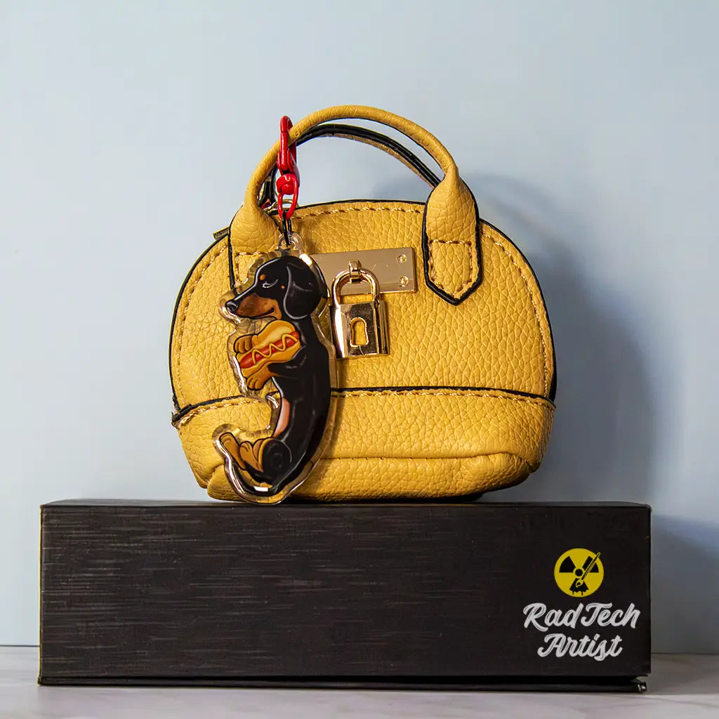 Acrylic Dachshund keychain with epoxy finish, featuring a black-and-tan dog hugging a hot dog with mustard. Attached with a bright red lobster clasp to a textured yellow mini purse with gold lock hardware, displayed on a black box with the Rad Tech Artist logo.