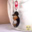 A die-cut acrylic keychain charm shaped like a black and tan dachshund holding a hot dog, attached to a red lobster clasp, shown hanging next to a white textured bag.