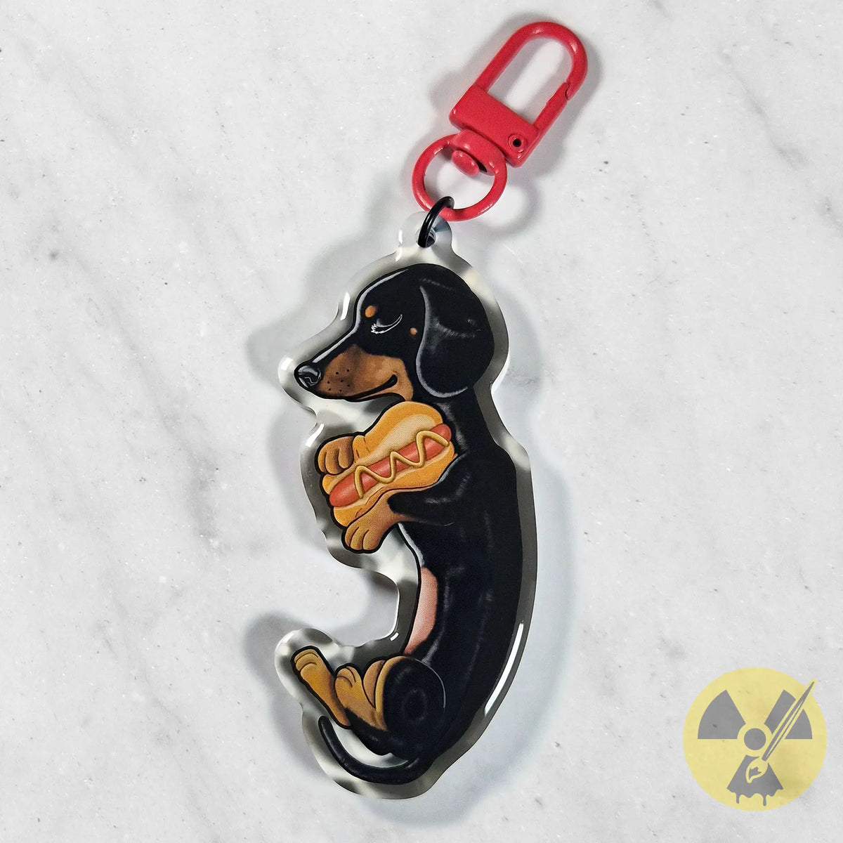 A dachshund-shaped acrylic keychain charm featuring a black and tan dog holding a hot dog, attached to a red metal swivel clasp, shown on a light marble surface.