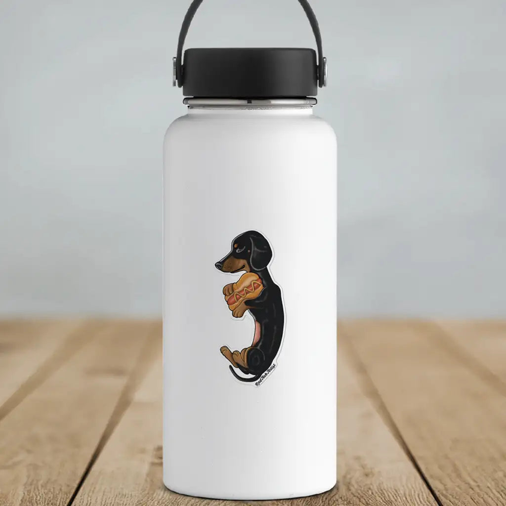 Mock-up image of a black and tan Dachshund vinyl sticker on a white water bottle. The sticker shows a side-view of a relaxed Dachshund sitting upright and holding a hot dog with mustard. The bottle rests on a wooden surface with a light background.