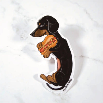 Sticker of a black and tan dachshund holding a hot dog on a marble surface