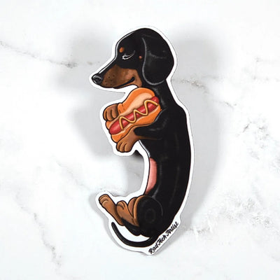 Magnet of a black and tan dachshund holding a hot dog on a marble background