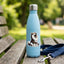 A light blue insulated water bottle with a silver cap stands on a wooden park bench, featuring a black-and-white husky dog sticker on the front. A navy blue dog leash with a silver clasp rests beside it, with a blurred green park background.