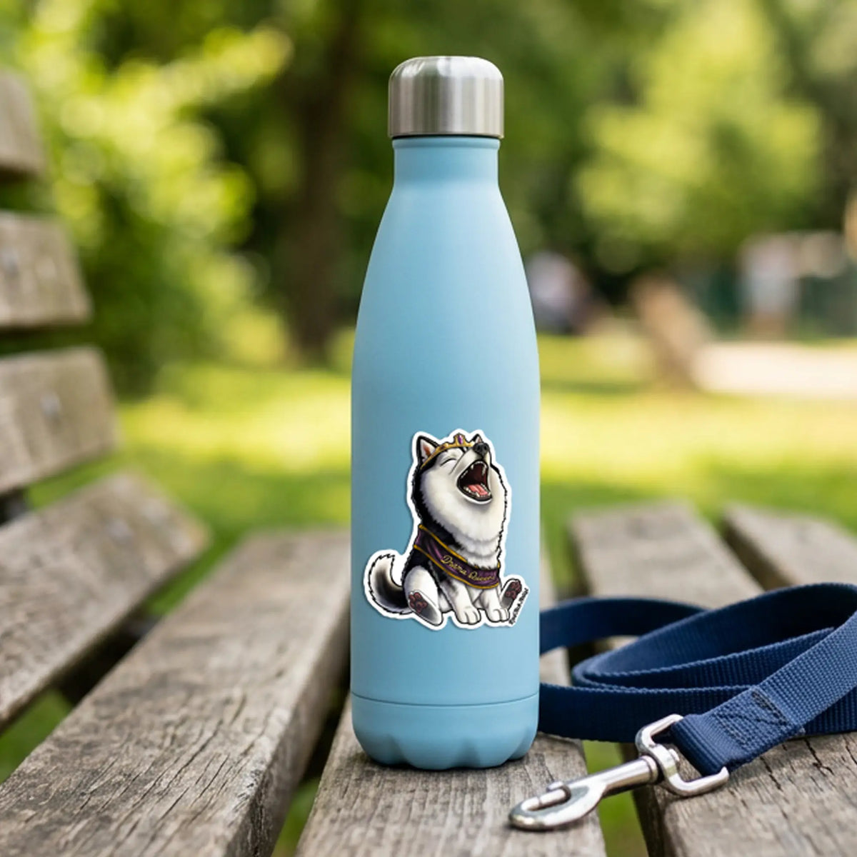 A light blue insulated water bottle with a silver cap stands on a wooden park bench, featuring a black-and-white husky dog sticker on the front. A navy blue dog leash with a silver clasp rests beside it, with a blurred green park background.