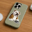An olive green phone case featuring a cartoon bulldog sticker sitting next to a dog bowl, shown on a wooden tabletop.