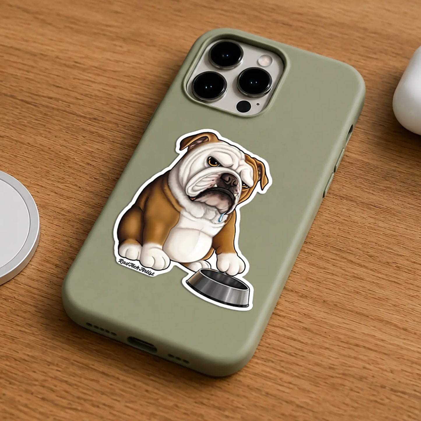An olive green phone case featuring a cartoon bulldog sticker sitting next to a dog bowl, shown on a wooden tabletop.