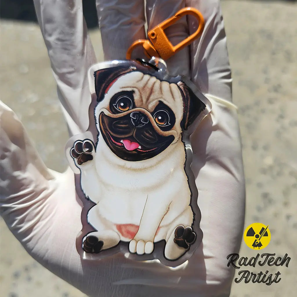 Fawn Pug acrylic keychain with epoxy finish, held in a gloved hand under sunlight. The pup is waving its paw with a happy expression, attached to an orange lobster clasp, with the Rad Tech Artist logo in the corner.