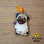 Acrylic fawn Pug keychain with epoxy finish, featuring a playful dog waving its paw. Attached to an orange lobster clasp, displayed on a warm wood background.