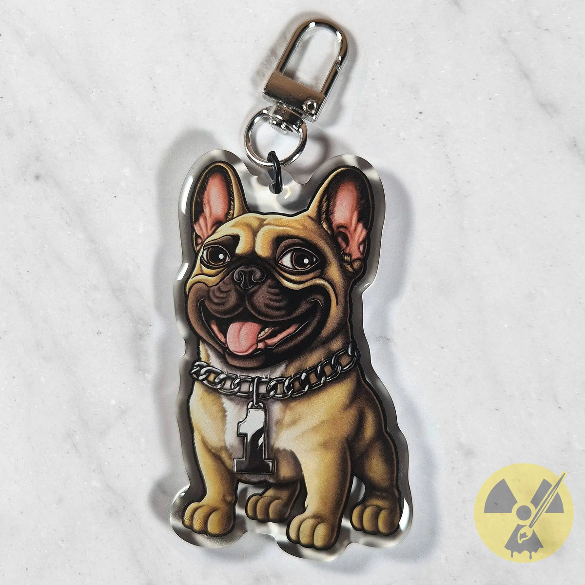 A die-cut acrylic keychain featuring a cartoon-style French Bulldog with a fawn coat, pink inner ears, and a silver chain collar, shown on a light marble background.