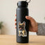 A hand holding a matte black insulated water bottle with a cartoon fawn French Bulldog sticker on the front, set against a softly blurred indoor background.