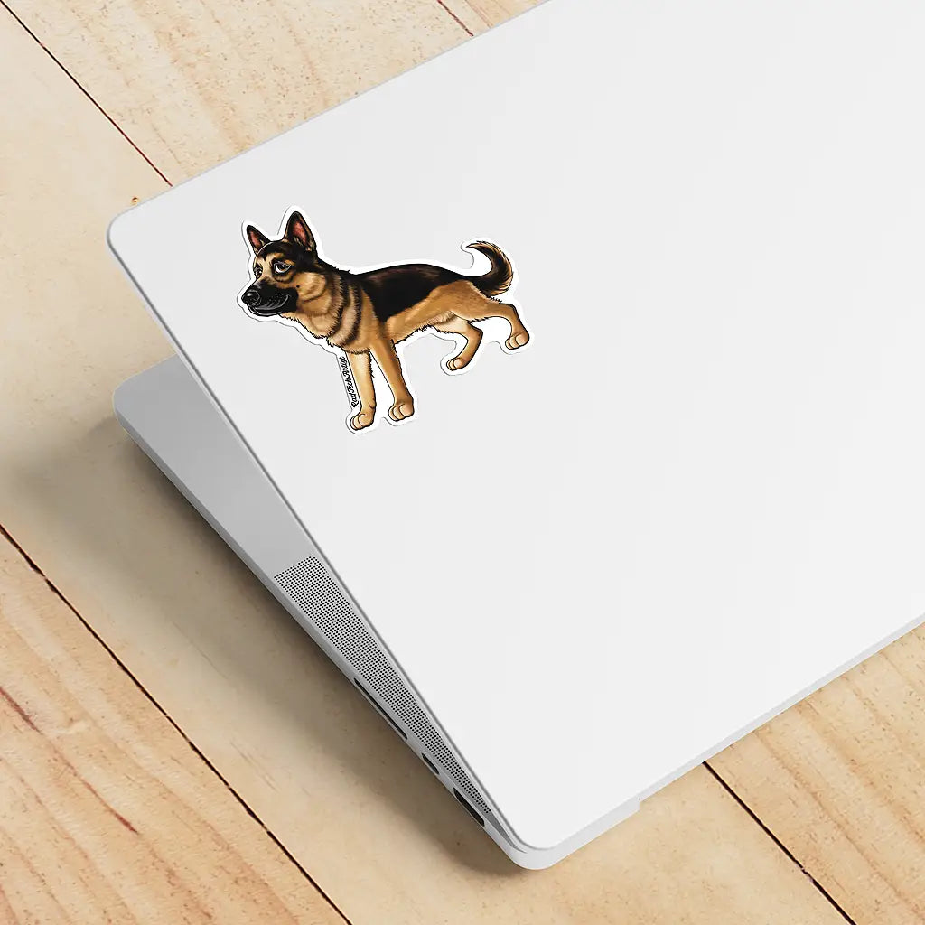 Mock-up image of a black and tan German Shepherd sticker placed on the back of a silver laptop. The dog stands alert with a confident stance and upright ears. The laptop rests on a light wood surface.