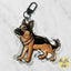 A die-cut acrylic keychain charm shaped like a German Shepherd dog, featuring a detailed illustration of the dog in black and tan, attached to a black metal lobster clasp, shown on a light marble background.