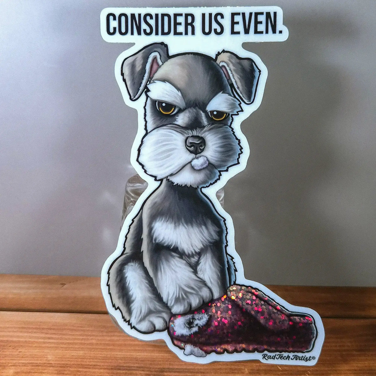 A die-cut sticker featuring an illustrated Schnauzer puppy dog with the text 'CONSIDER US EVEN.' above it, displayed standing on a wooden surface against a gray background.