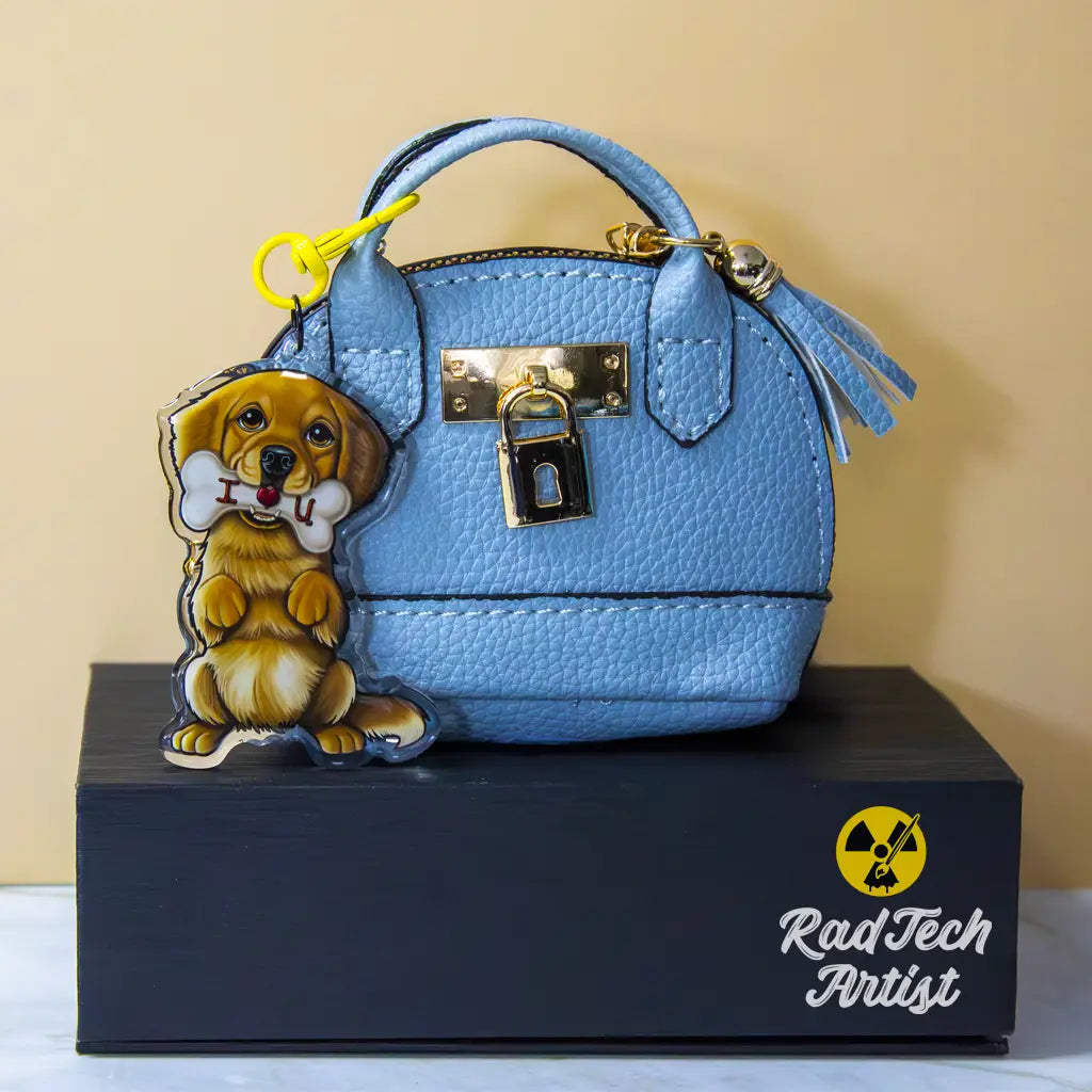 Golden Retriever acrylic keychain with epoxy finish, featuring a fluffy golden pup holding a white bone with “I ❤️ U” text. Attached with a yellow lobster clasp to a pastel blue mini purse with gold hardware, displayed on a black box with the Rad Tech Artist logo.