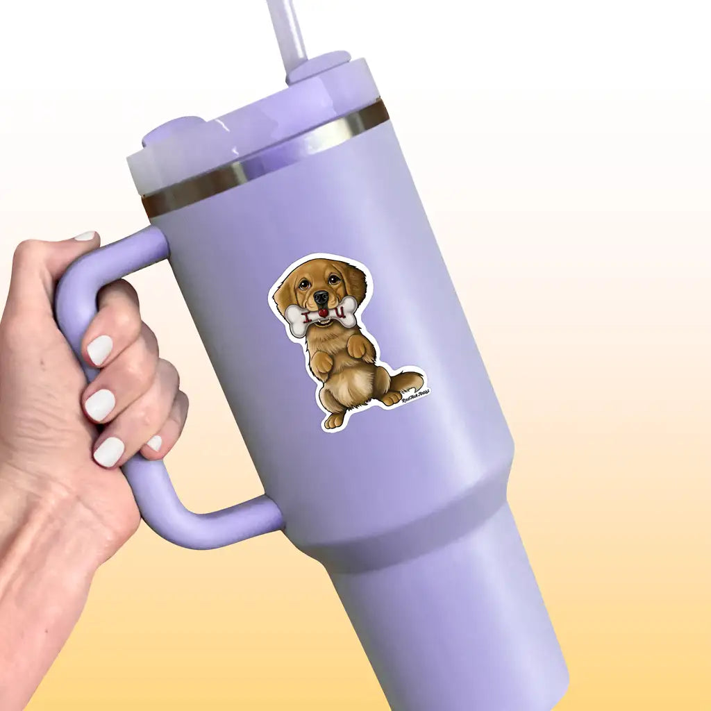 Mock-up image of a Golden Retriever vinyl sticker on a lavender tumbler cup. The sticker shows a happy golden puppy sitting upright with a large bone in its mouth. The tumbler is held by a hand with pale nail polish, set against a soft yellow background.