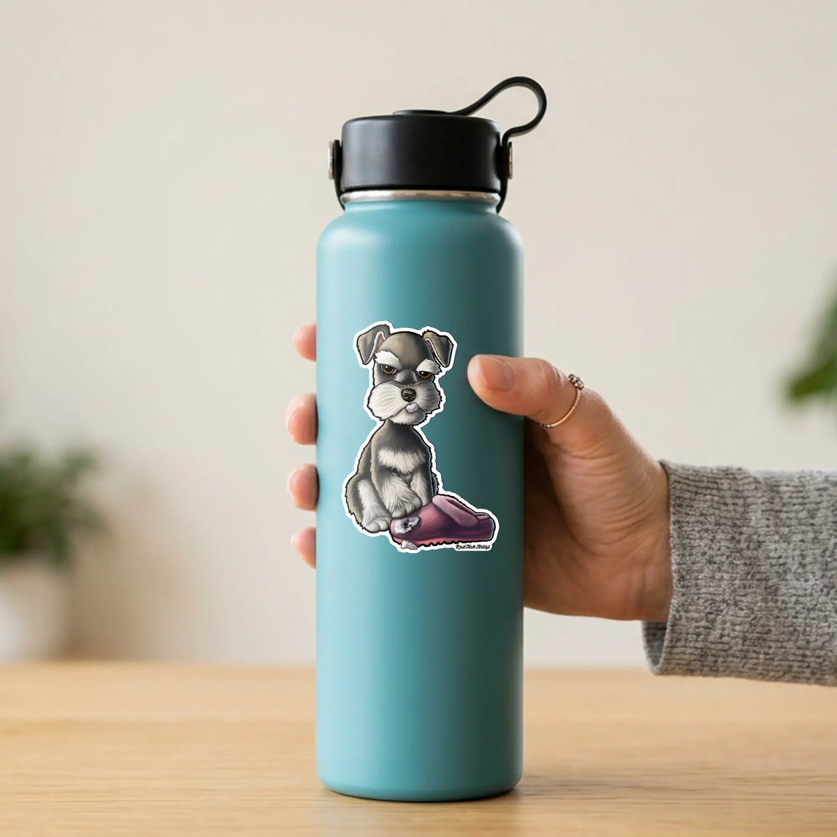 A hand holding a teal insulated water bottle with a black lid, featuring a cartoon Schnauzer sticker on the front, set against a softly blurred indoor background.