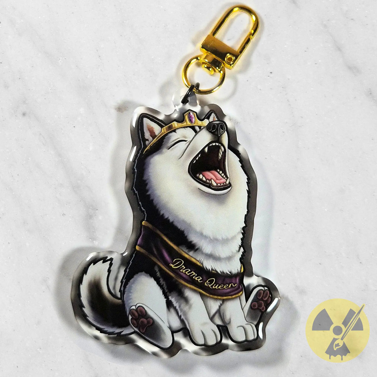 A die-cut acrylic keychain charm featuring an illustrated whining husky dog wearing a purple sash labeled 'Drama Queen' with a gold clasp, photographed on a light marble-like surface.