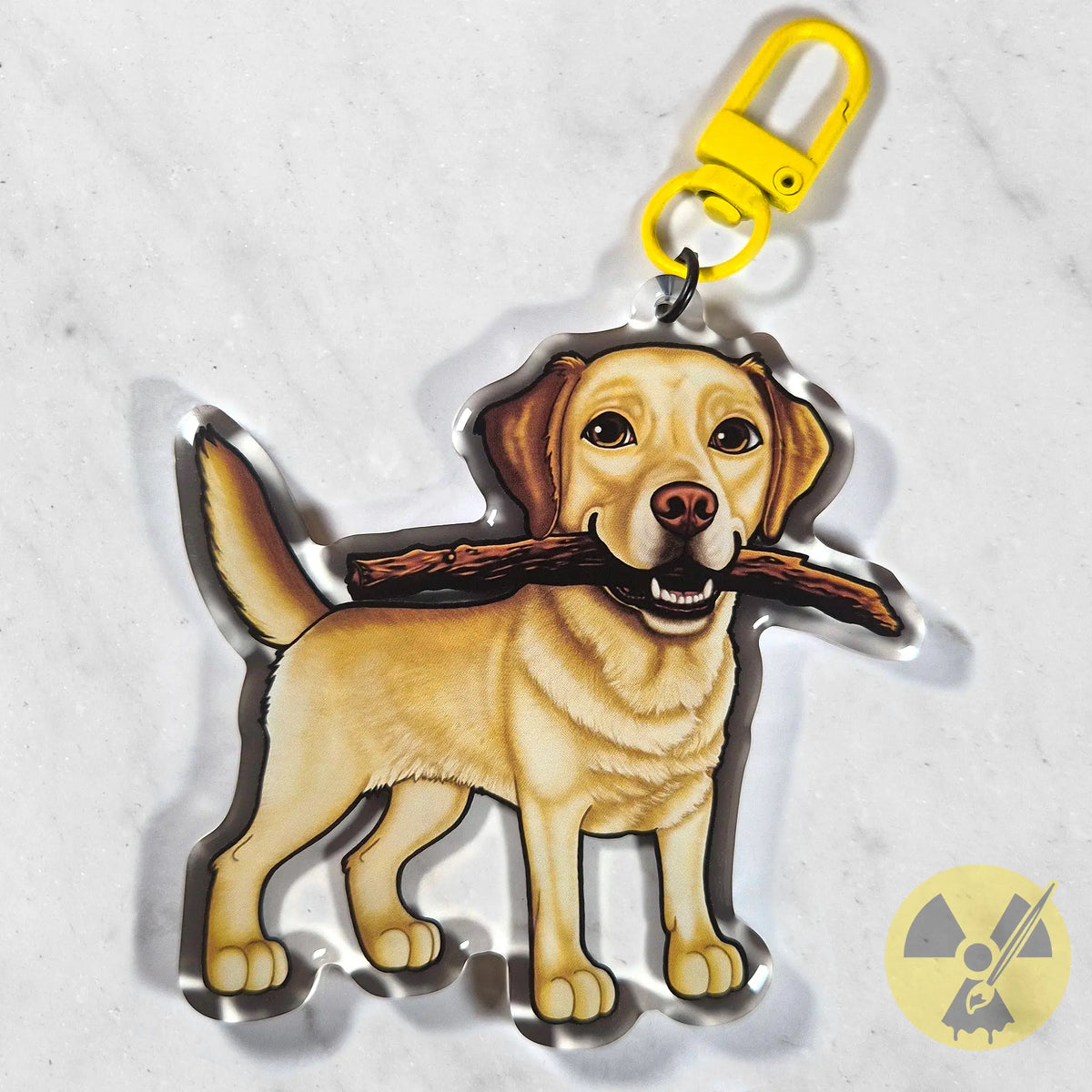 A die-cut acrylic keychain featuring a cartoon-style yellow Labrador retriever holding a brown stick in its mouth, attached to a bright yellow swivel clasp, shown on a light marble background.