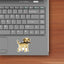 Mock-up image of a yellow Labrador Retriever sticker placed on a silver laptop. The Lab is smiling with a large stick held in its mouth, standing with a friendly, alert posture. The laptop is shown on a wooden desk next to a keyboard.