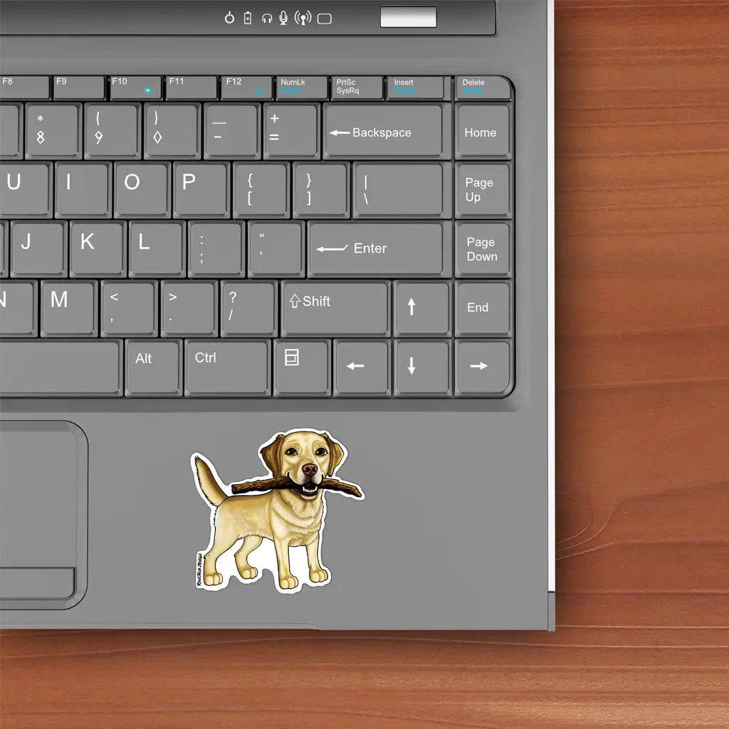 Mock-up image of a yellow Labrador Retriever sticker placed on a silver laptop. The Lab is smiling with a large stick held in its mouth, standing with a friendly, alert posture. The laptop is shown on a wooden desk next to a keyboard.